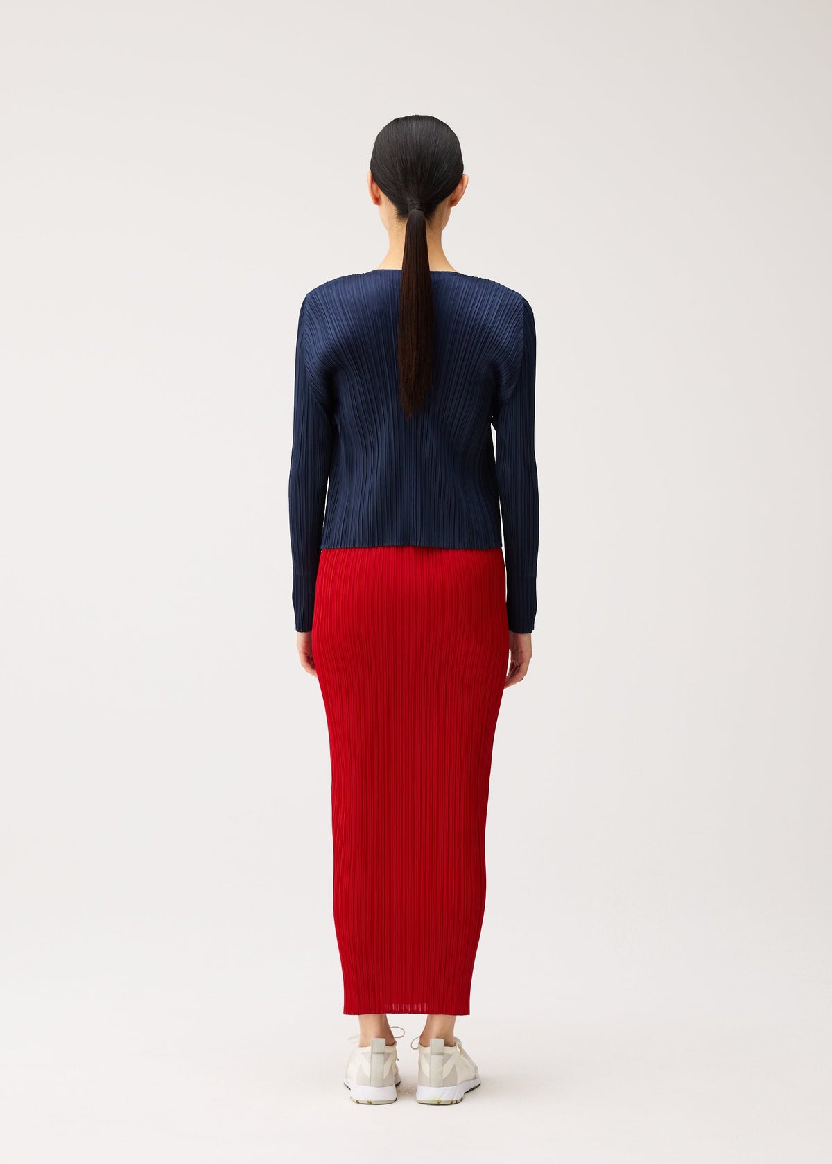 Seen from behind, a person wears a PLEATS PLEASE ISSEY MIYAKE NEW COLORFUL BASICS 5 navy blue long-sleeved pleated top and a bright red ankle-length pleated skirt, paired with white sneakers. view 9 of 10