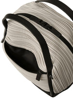 A close-up of the BIAS PLEATS BAG by PLEATS PLEASE ISSEY MIYAKE shows its beige, ribbed pleats, black handles, and partially open black zipper revealing a black interior. The shoulder bag comes with an attached strap for versatile carrying.