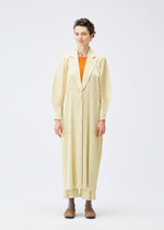 A person stands facing forward in a cream pleated MONTHLY COLORS : DECEMBER coat and pants by PLEATS PLEASE ISSEY MIYAKE, with an orange top and brown shoes, set against a plain white background.