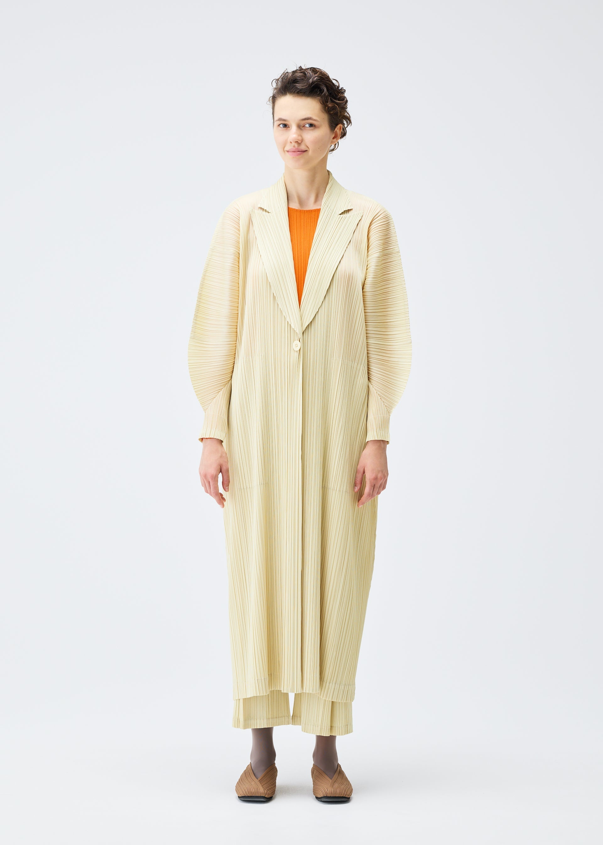 Zoomed view A person stands facing forward in a cream pleated MONTHLY COLORS : DECEMBER coat and pants by PLEATS PLEASE ISSEY MIYAKE, with an orange top and brown shoes, set against a plain white background.