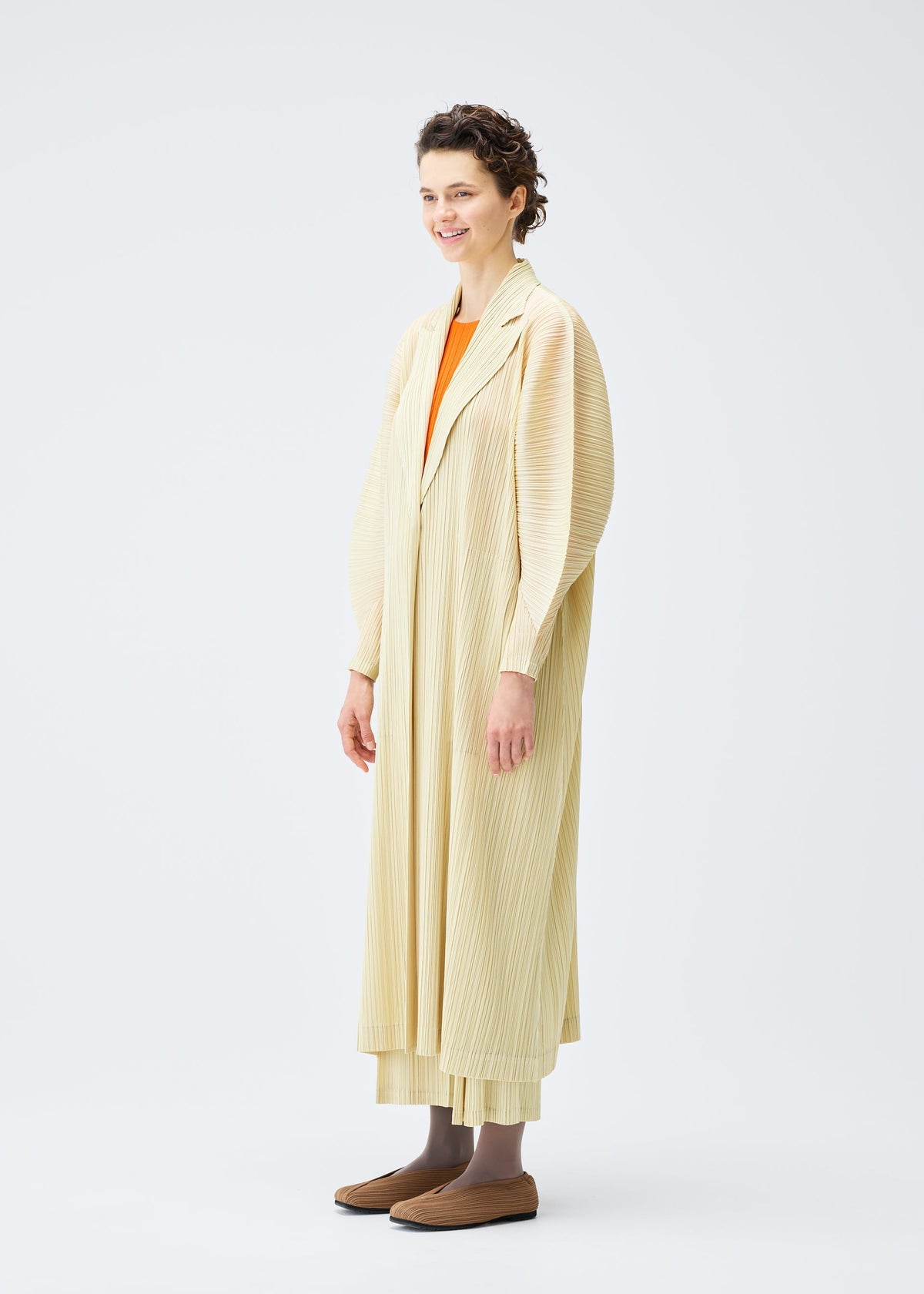 A person stands against a plain white background, smiling and looking slightly to the side, wearing the MONTHLY COLORS : DECEMBER set by PLEATS PLEASE ISSEY MIYAKE—a collared long cardigan with dolman sleeves over matching pants. view 7 of 10