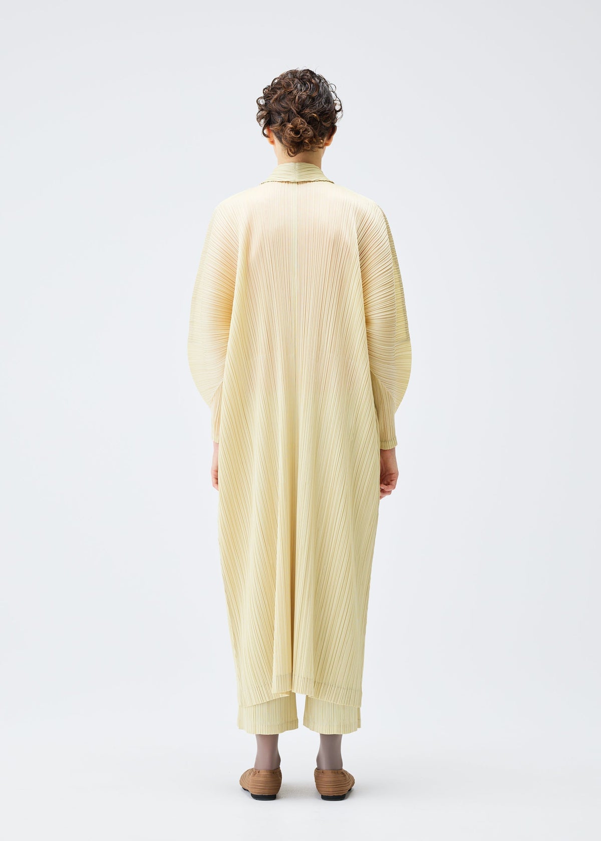 A person with short curly hair faces away, wearing the MONTHLY COLORS : DECEMBER pleated robe and matching pants by PLEATS PLEASE ISSEY MIYAKE, paired with brown shoes, set against a plain white background. view 8 of 10