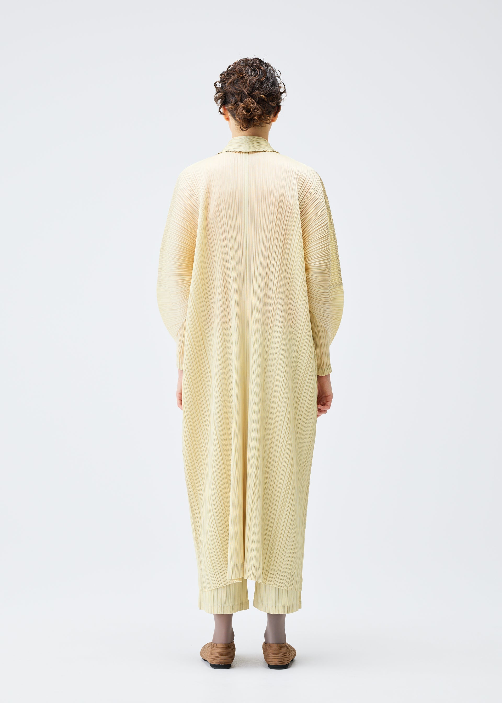 Zoomed view A person with short curly hair faces away, wearing the MONTHLY COLORS : DECEMBER pleated robe and matching pants by PLEATS PLEASE ISSEY MIYAKE, paired with brown shoes, set against a plain white background.