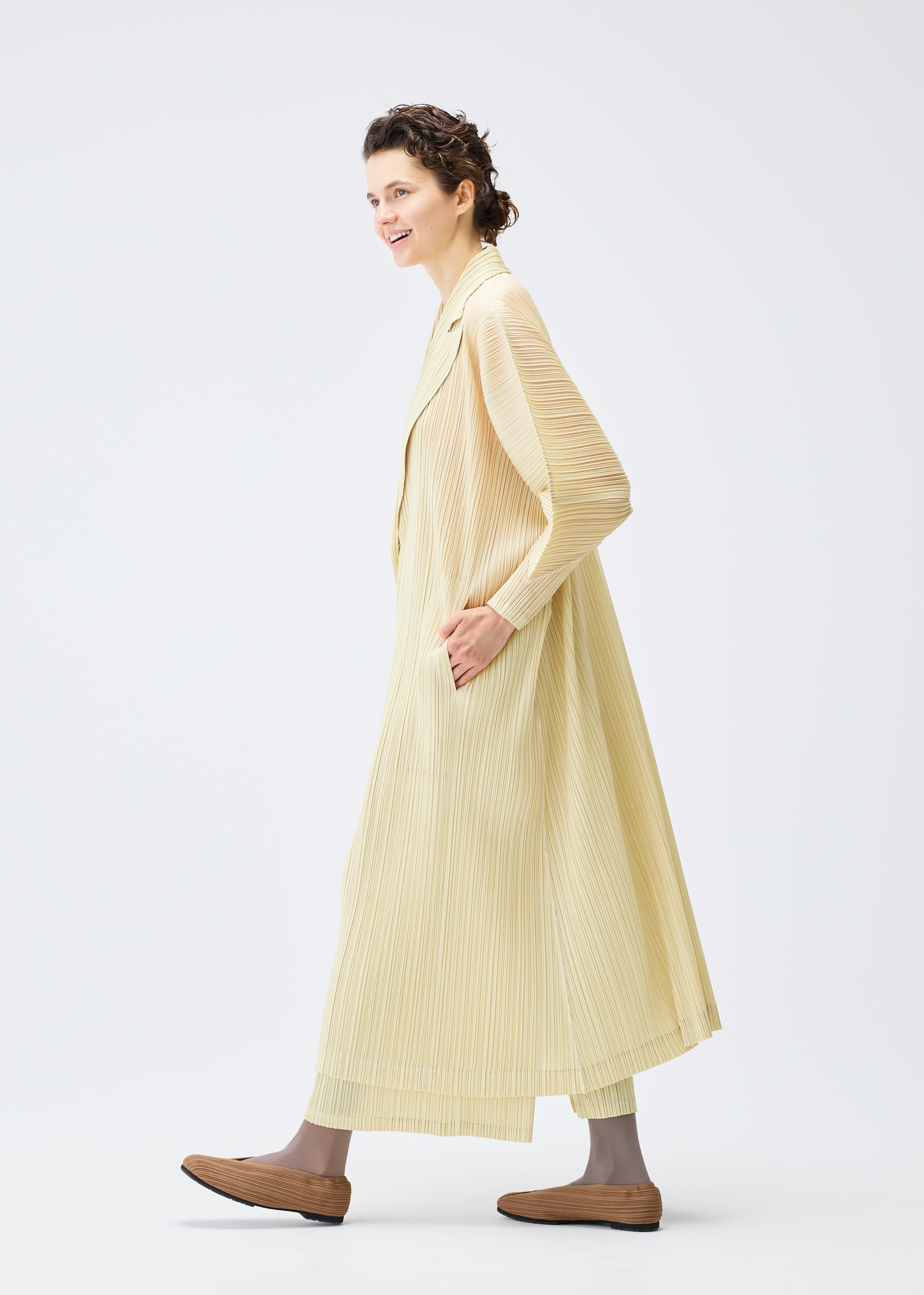 Zoomed view A smiling person with short, wavy hair models the light yellow MONTHLY COLORS : DECEMBER long cardigan and matching wide-leg pants from PLEATS PLEASE ISSEY MIYAKE, standing against a plain white background.