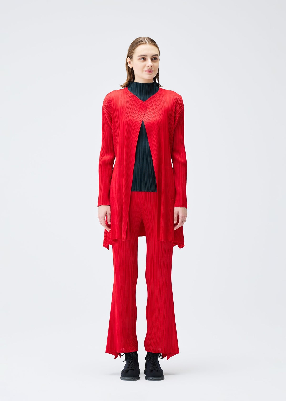 A woman faces forward in the OLE! pleated long red cardigan and matching pants by PLEATS PLEASE ISSEY MIYAKE over a black turtleneck, paired with black lace-up shoes, set against a plain white background. view 4 of 9