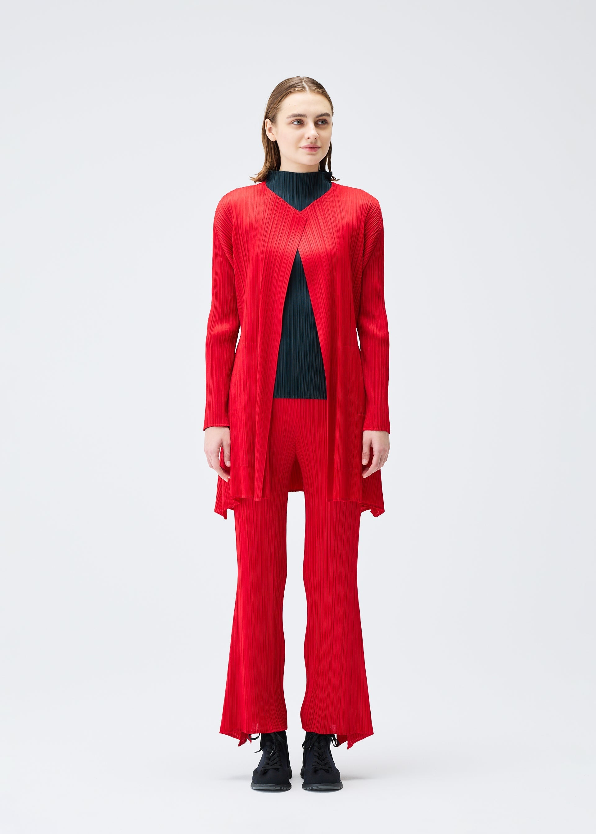 Zoomed view A woman faces forward in the OLE! pleated long red cardigan and matching pants by PLEATS PLEASE ISSEY MIYAKE over a black turtleneck, paired with black lace-up shoes, set against a plain white background.