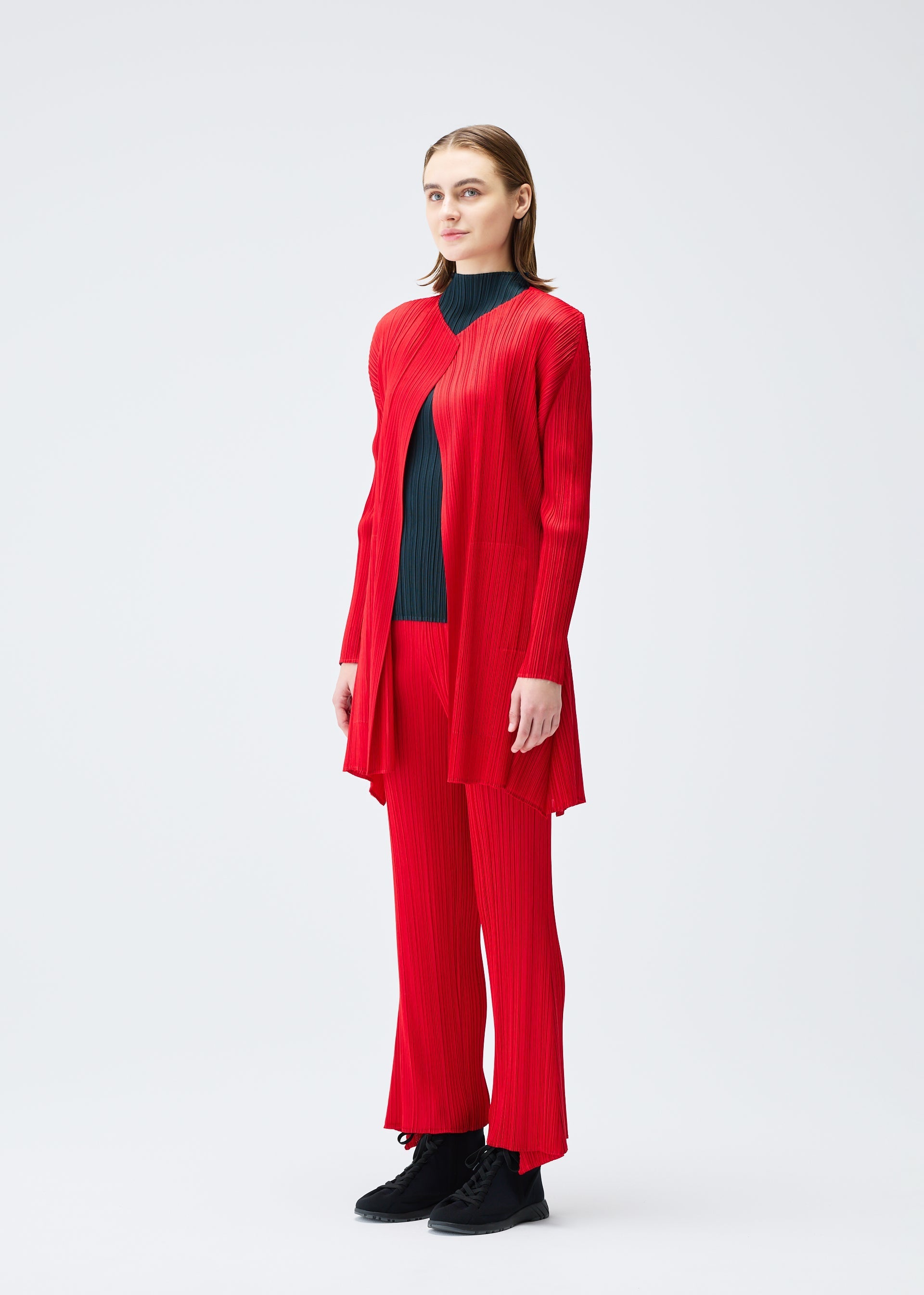 Zoomed view A person with straight, shoulder-length hair and a neutral expression wears the bright red OLE! set from PLEATS PLEASE ISSEY MIYAKE—featuring a flared long cardigan and matching wide-leg pants over a dark top, styled with black shoes against a white background.