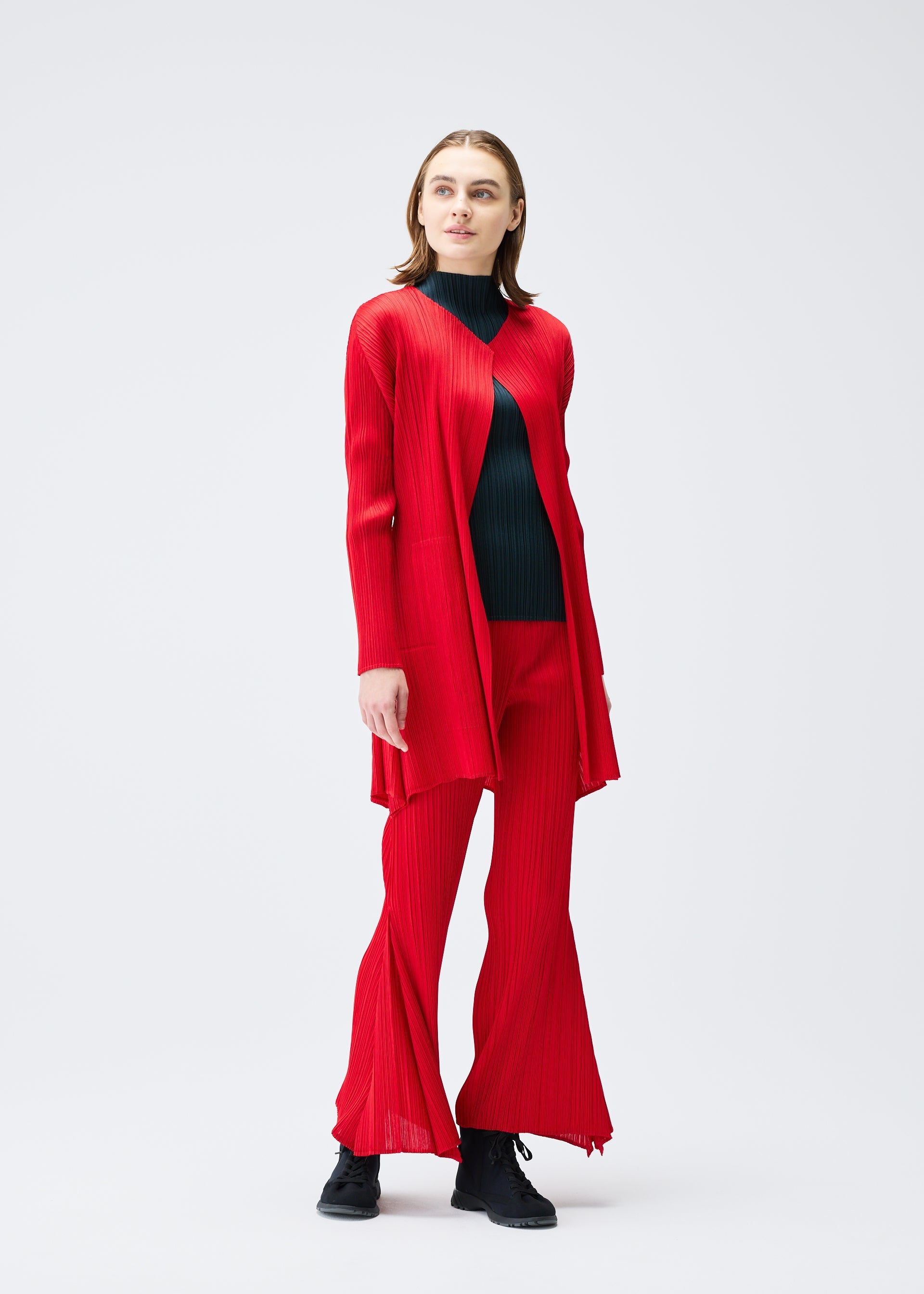 Zoomed view A person with straight, shoulder-length brown hair stands against a plain light background, wearing the OLE! set by PLEATS PLEASE ISSEY MIYAKE—long red cardigan and matching flared pleated pants—plus a black ribbed turtleneck and black shoes.