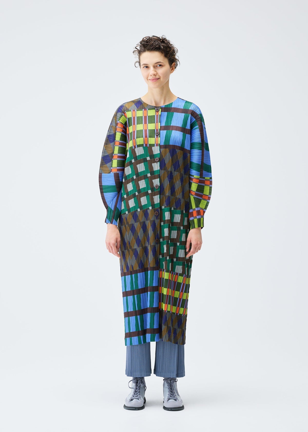 A person with short curly hair and a neutral expression wears the HARMONY coat by PLEATS PLEASE ISSEY MIYAKE, featuring colorful geometric patterns over blue pleated pants and gray shoes against a white background. view 4 of 7