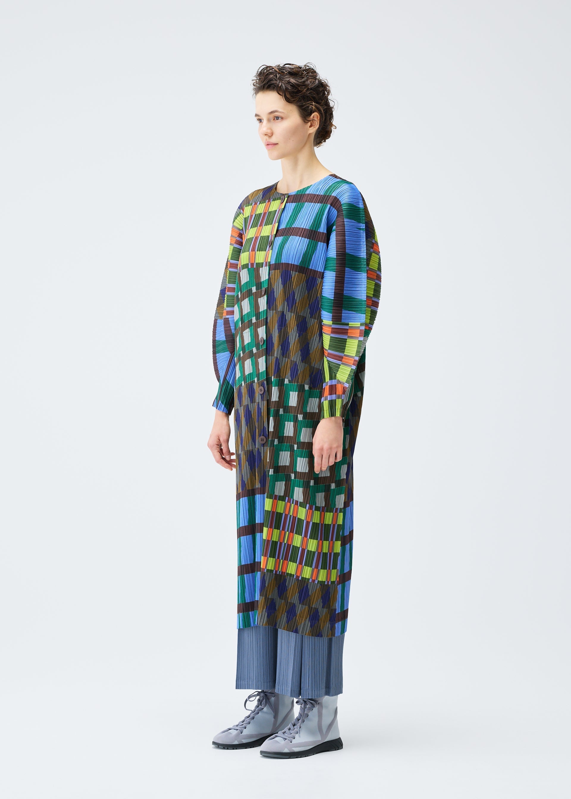 Zoomed view A person stands in profile against a plain white background, wearing the HARMONY coat by PLEATS PLEASE ISSEY MIYAKE—a colorful, geometric print piece styled over blue pleated pants and paired with silver lace-up boots.