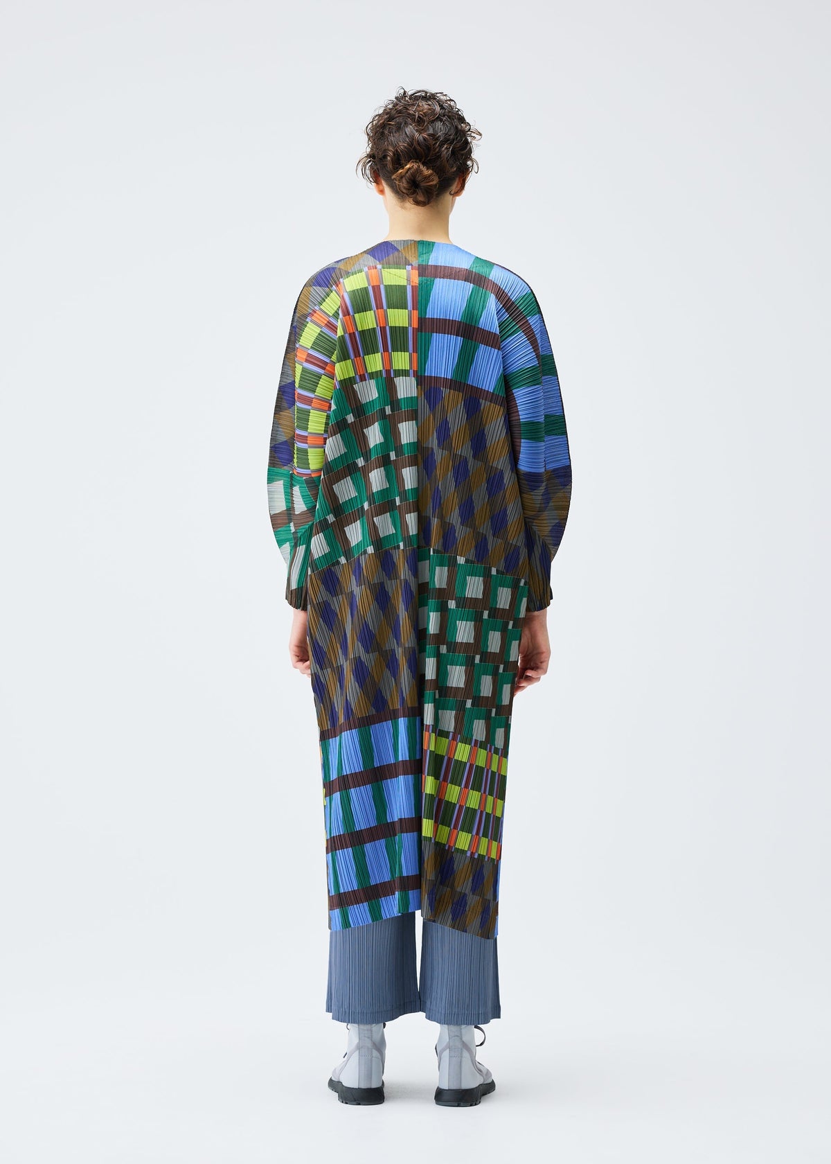 A person faces away, wearing the HARMONY coat by PLEATS PLEASE ISSEY MIYAKE, featuring a bold geometric print in blue, green, yellow, and red over pleated blue pants and gray shoes against a white background. view 6 of 7