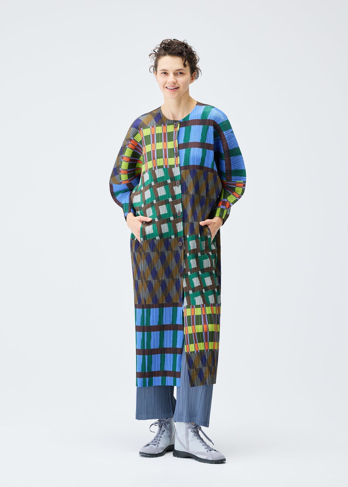 A person with short curly hair models the HARMONY coat by PLEATS PLEASE ISSEY MIYAKE—featuring a colorful geometric check print—paired with blue pleated pants and silver lace-up shoes, against a plain white background. view 7 of 7