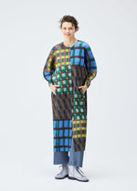 A person with short curly hair models the HARMONY coat by PLEATS PLEASE ISSEY MIYAKE—featuring a colorful geometric check print—paired with blue pleated pants and silver lace-up shoes, against a plain white background.