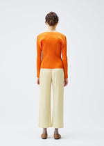 A person with short curly hair faces away, wearing the MONTHLY COLORS : DECEMBER orange ribbed long-sleeve top and off-white pleated wide-leg pants with pockets by PLEATS PLEASE ISSEY MIYAKE, paired with brown shoes, against a plain background.