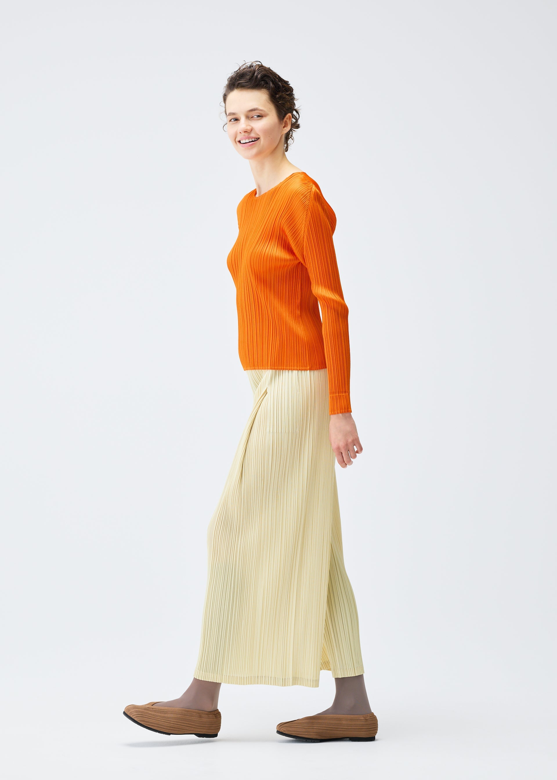 Zoomed view A person smiles and walks sideways against a light background, wearing PLEATS PLEASE ISSEY MIYAKE's MONTHLY COLORS : DECEMBER—a vibrant orange long-sleeve top with tucks, a cream pleated skirt, brown shoes, and taupe socks.
