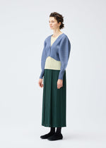 A woman with short, curly hair wears the MONTHLY COLORS : DECEMBER pleated skirt from PLEATS PLEASE ISSEY MIYAKE, paired with a light yellow top and ribbed blue cardigan. She stands against a white background, exuding subtle joy in her stance.