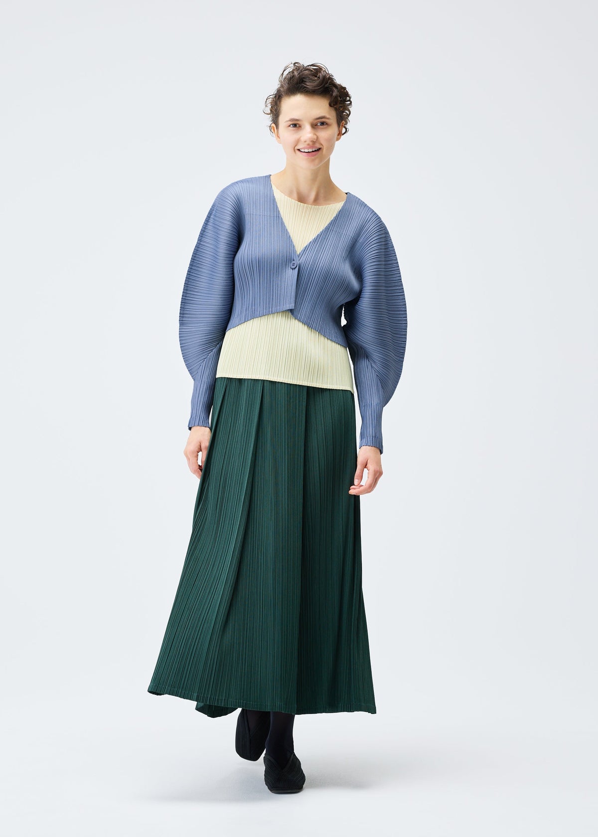 A person smiles gently against a white background, wearing the PLEATS PLEASE ISSEY MIYAKE MONTHLY COLORS : DECEMBER collection, featuring a light blue textured cardigan, pale yellow top, long dark green skirt, and black shoes. view 9 of 10