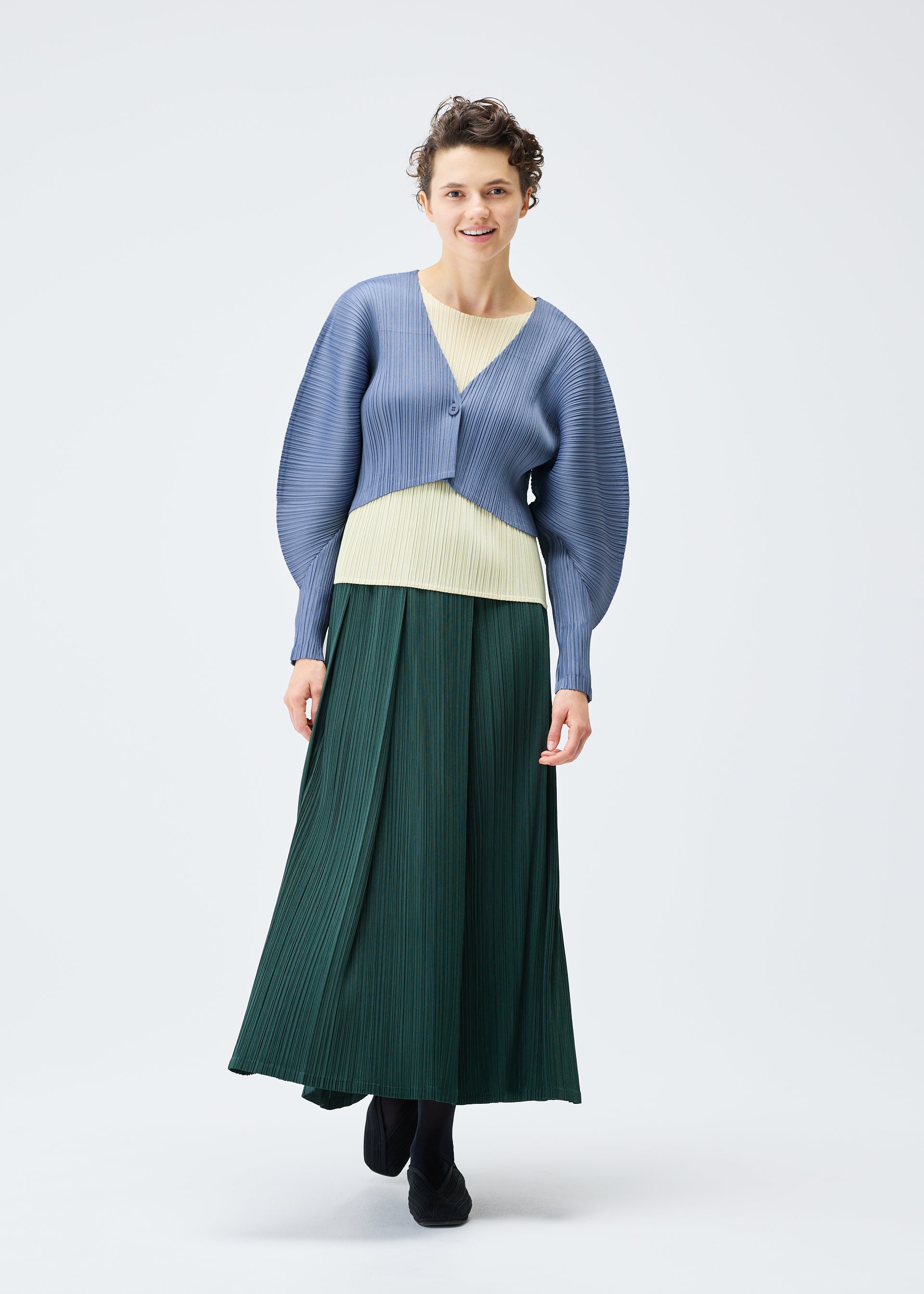 Zoomed view A person smiles gently against a white background, wearing the PLEATS PLEASE ISSEY MIYAKE MONTHLY COLORS : DECEMBER collection, featuring a light blue textured cardigan, pale yellow top, long dark green skirt, and black shoes.