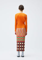 A person stands facing away in the HARMONY long-sleeved orange pleated top and colorful geometric print skirt with pockets by PLEATS PLEASE ISSEY MIYAKE, paired with light gray ankle boots against a plain white background.