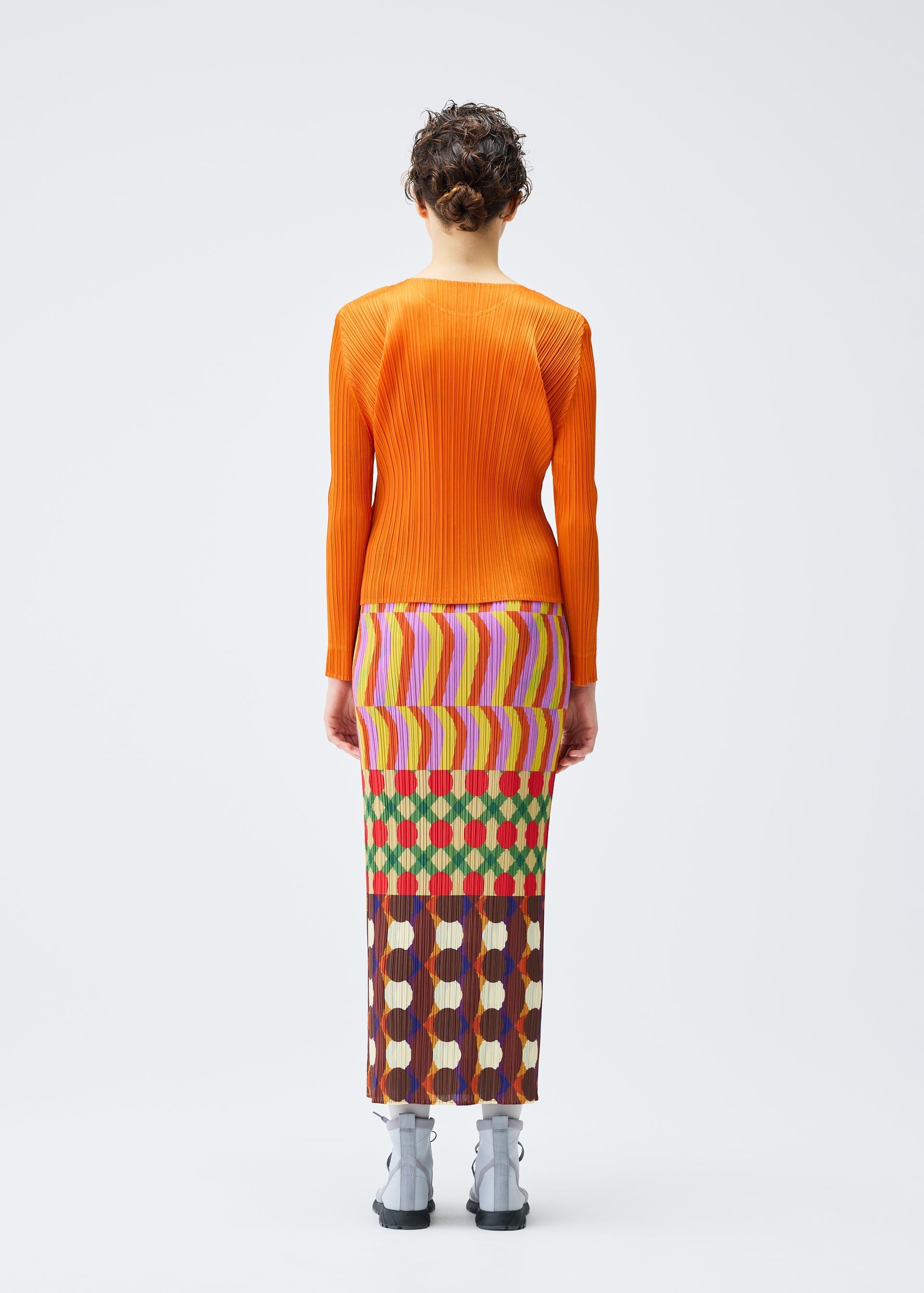 Zoomed view A person stands facing away in the HARMONY long-sleeved orange pleated top and colorful geometric print skirt with pockets by PLEATS PLEASE ISSEY MIYAKE, paired with light gray ankle boots against a plain white background.