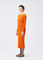 A woman with short curly hair stands sideways, smiling in the MONTHLY COLORS : DECEMBER long-sleeved, ankle-length orange ribbed dress by PLEATS PLEASE ISSEY MIYAKE, styled with gray tights and brown flats against a white background.