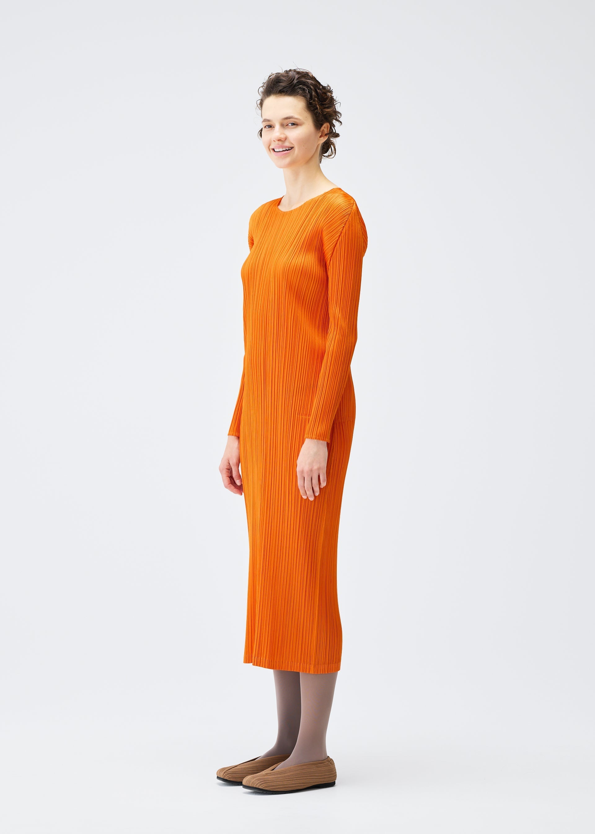 Zoomed view A woman with short curly hair stands sideways, smiling in the MONTHLY COLORS : DECEMBER long-sleeved, ankle-length orange ribbed dress by PLEATS PLEASE ISSEY MIYAKE, styled with gray tights and brown flats against a white background.