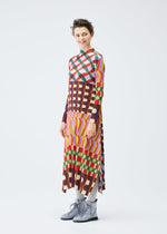 A person with short hair smiles while wearing the HARMONY dress by PLEATS PLEASE ISSEY MIYAKE, featuring a long, colorful geometric print with stripes, paired with gray tights and light gray lace-up boots against a white background.