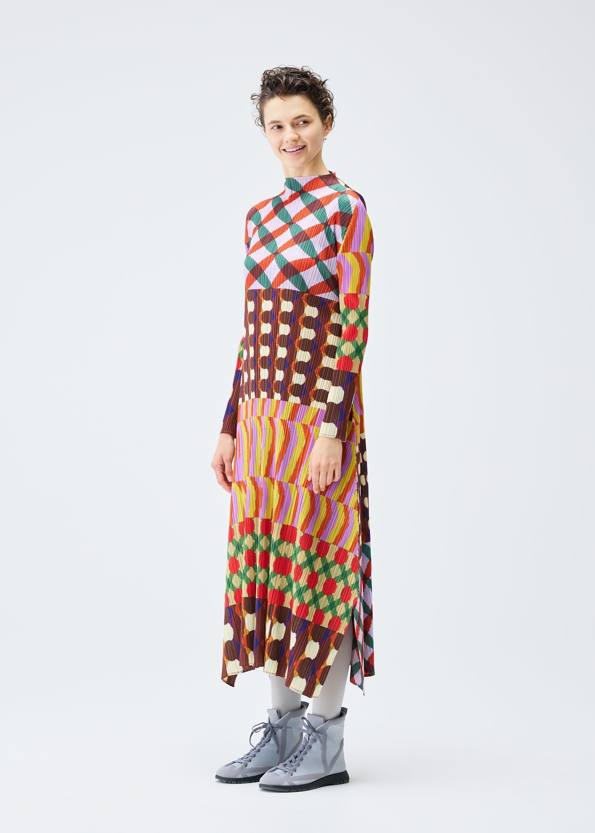 Zoomed view A person with short hair smiles while wearing the HARMONY dress by PLEATS PLEASE ISSEY MIYAKE, featuring a long, colorful geometric print with stripes, paired with gray tights and light gray lace-up boots against a white background.