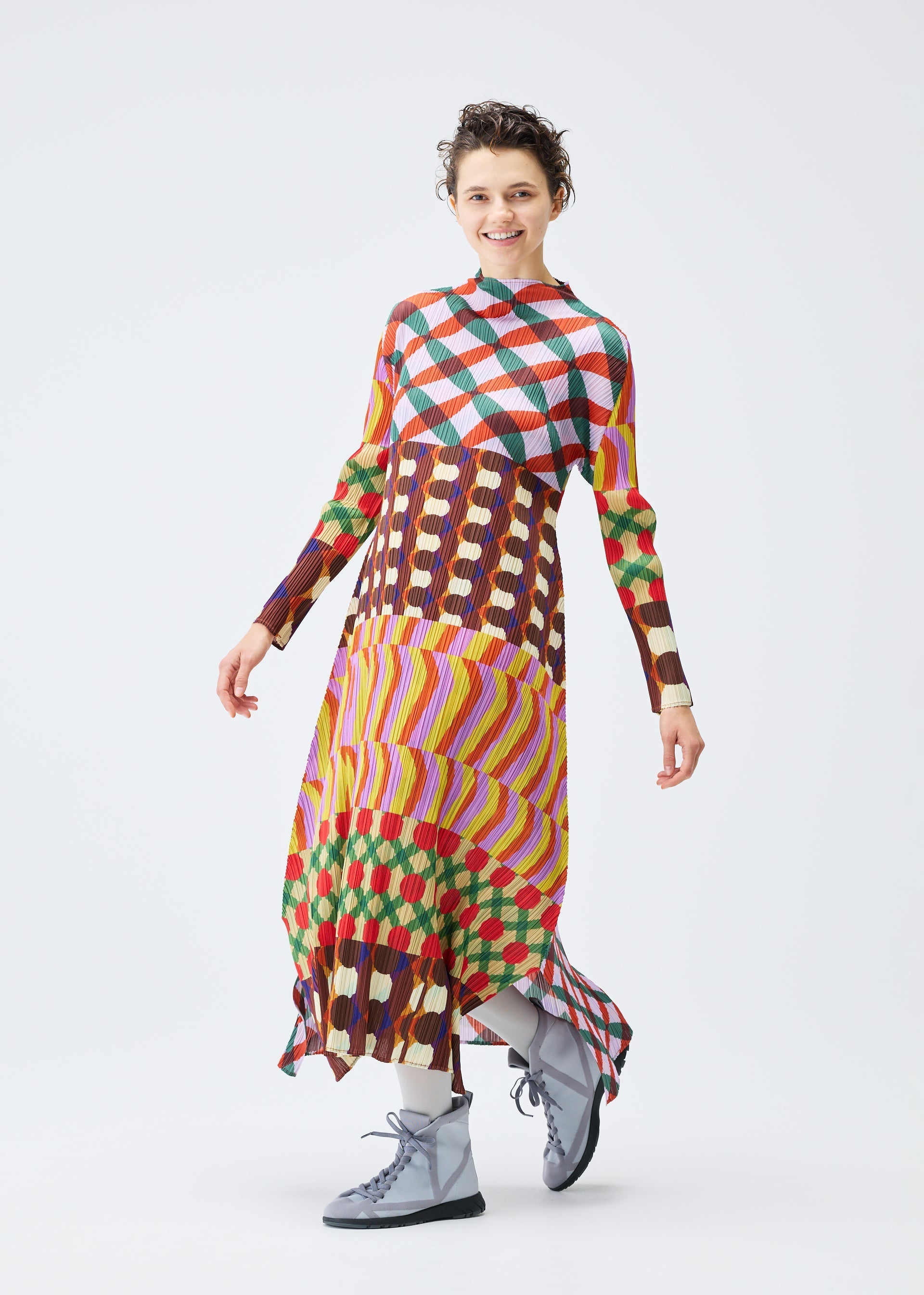 Zoomed view A person stands smiling in the HARMONY dress by PLEATS PLEASE ISSEY MIYAKE, featuring a long, colorful geometric print and long sleeves, paired with gray lace-up boots against a plain white background.