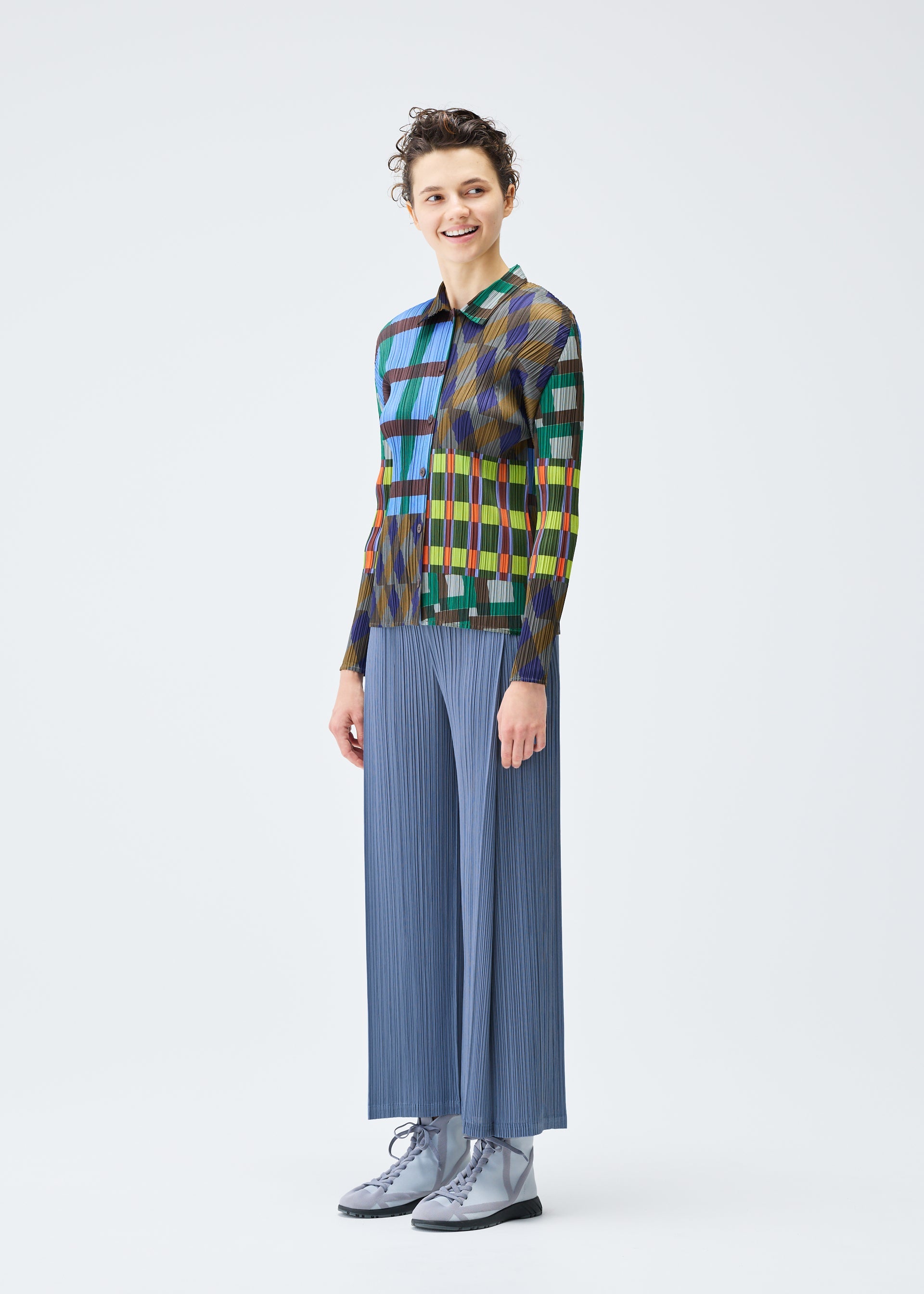 Zoomed view A person smiles in the HARMONY long-sleeved geometric print shirt and wide blue pleated pants by PLEATS PLEASE ISSEY MIYAKE, paired with light gray lace-up boots, against a plain white background.