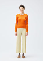 A person stands smiling in the MONTHLY COLORS : DECEMBER collection by PLEATS PLEASE ISSEY MIYAKE, featuring a bright orange long-sleeved top and loose cream pleated pants. The joyful colors pop against a plain white background.