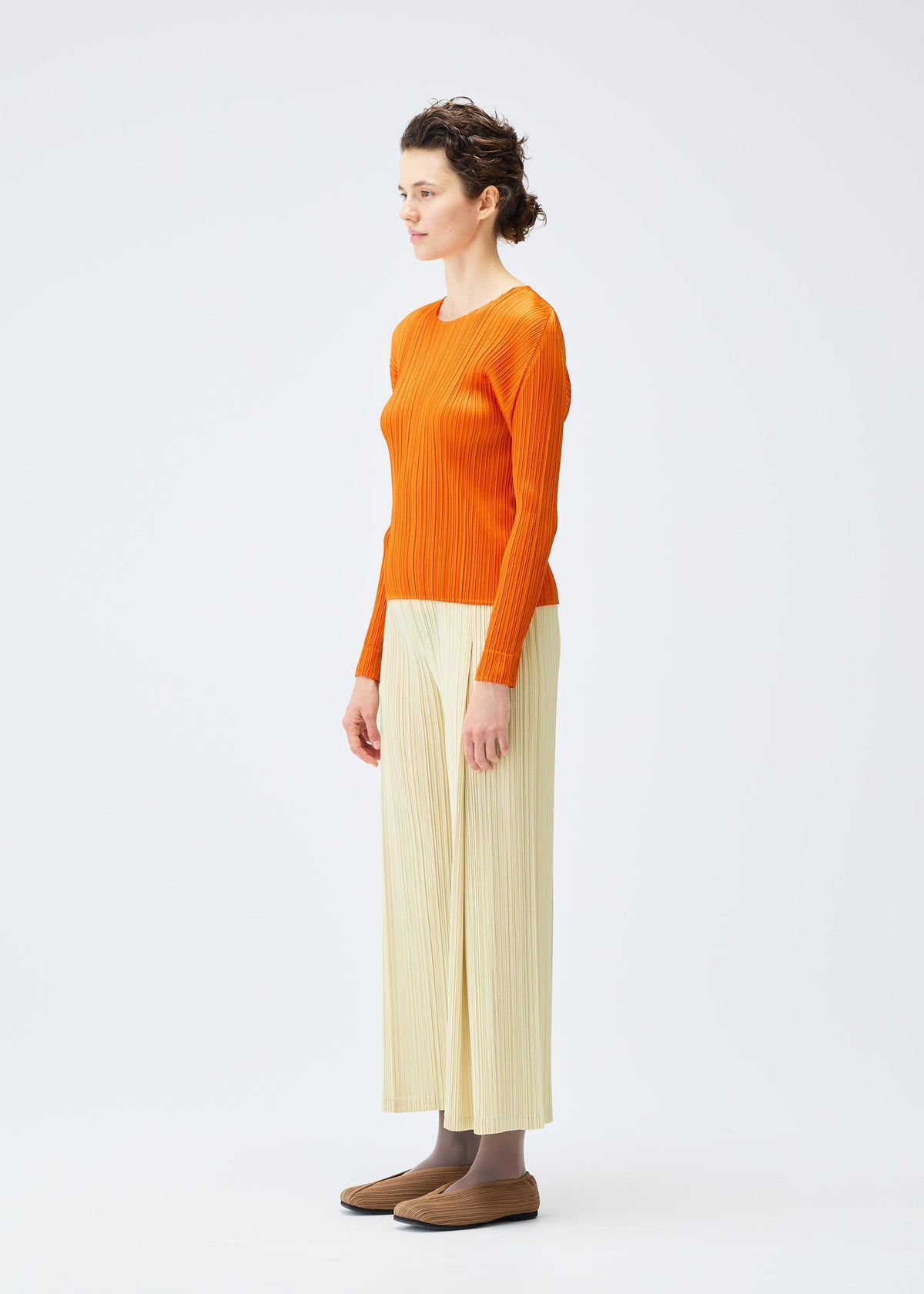 A person stands sideways against a white background, wearing the MONTHLY COLORS : DECEMBER orange top and cream pleated wide-leg pants by PLEATS PLEASE ISSEY MIYAKE, paired with brown shoes and gray socks. view 8 of 10