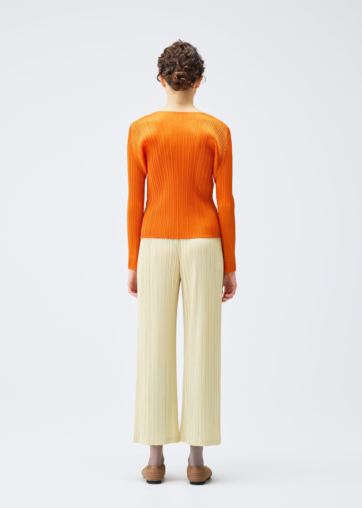 A person with short curly hair faces away, wearing the PLEATS PLEASE ISSEY MIYAKE MONTHLY COLORS : DECEMBER orange round-neck long-sleeved top and light beige pleated pants, standing against a plain white background. view 9 of 10