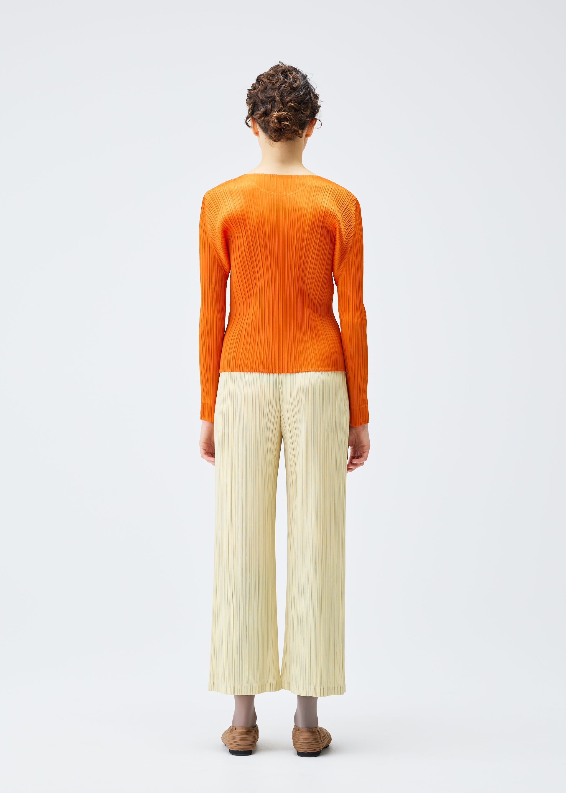 Zoomed view A person with short curly hair faces away, wearing the PLEATS PLEASE ISSEY MIYAKE MONTHLY COLORS : DECEMBER orange round-neck long-sleeved top and light beige pleated pants, standing against a plain white background.
