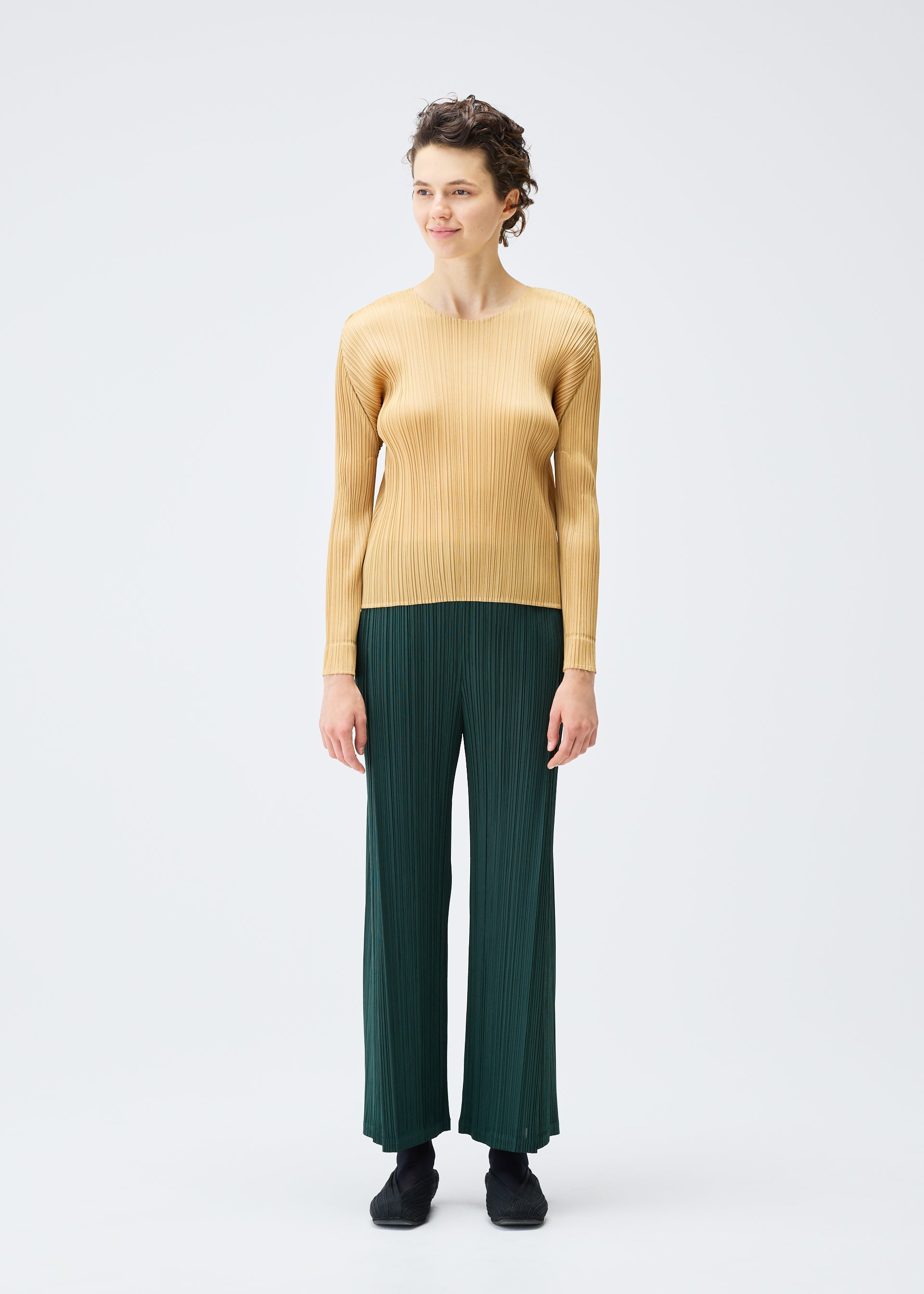 Zoomed view A person models the MONTHLY COLORS : DECEMBER set by PLEATS PLEASE ISSEY MIYAKE—a long-sleeved, round-neck light tan top with dark green pleated pants—against a white backdrop, looking slightly to the side with a faint smile.