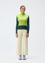 Against a plain white background, a person in the PLEATS PLEASE ISSEY MIYAKE TUNE top pairs its green and yellow layers with wide cream pants and gray sneakers. Their short, braided hair complements the modern ensemble.