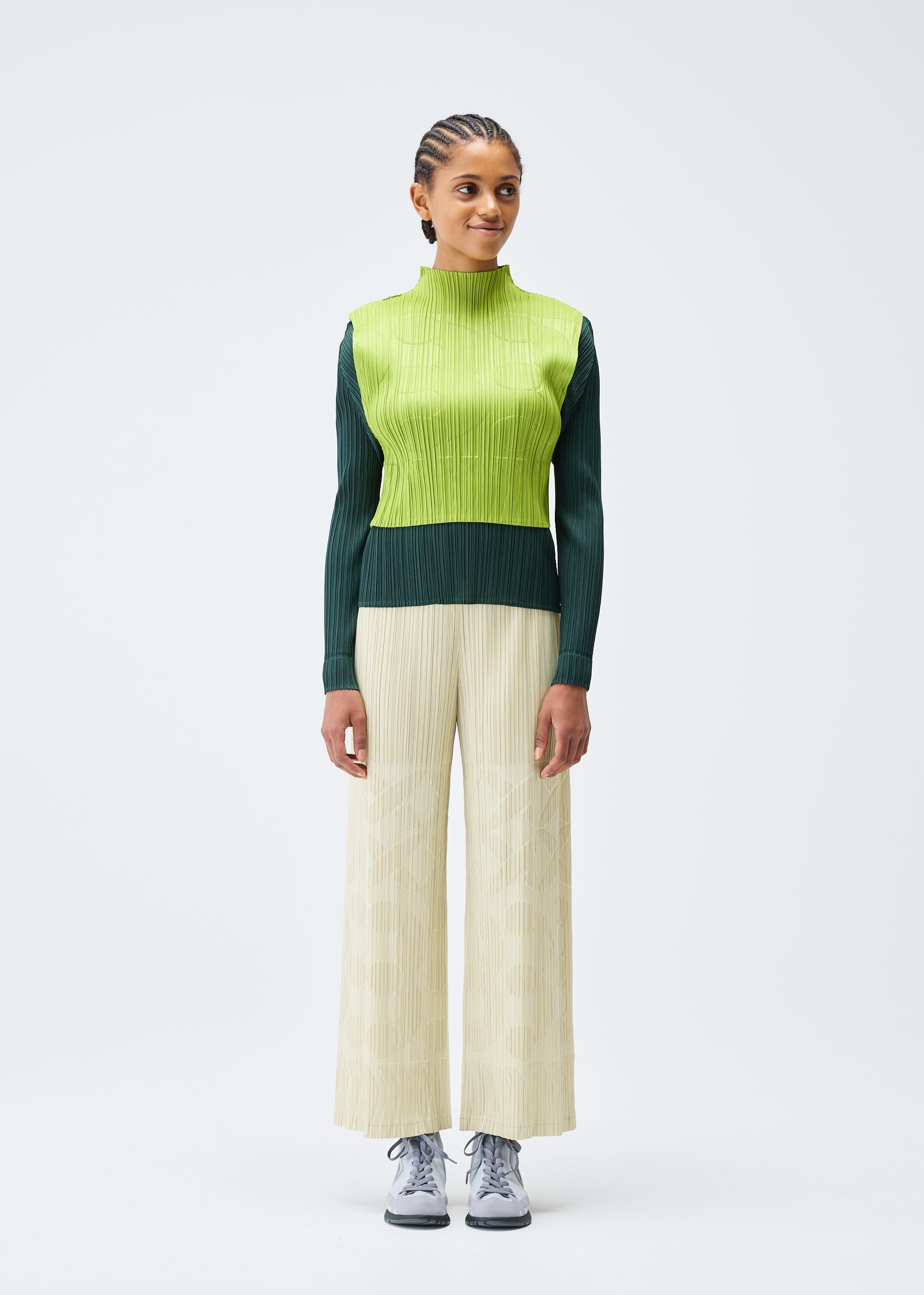 Zoomed view Against a plain white background, a person in the PLEATS PLEASE ISSEY MIYAKE TUNE top pairs its green and yellow layers with wide cream pants and gray sneakers. Their short, braided hair complements the modern ensemble.