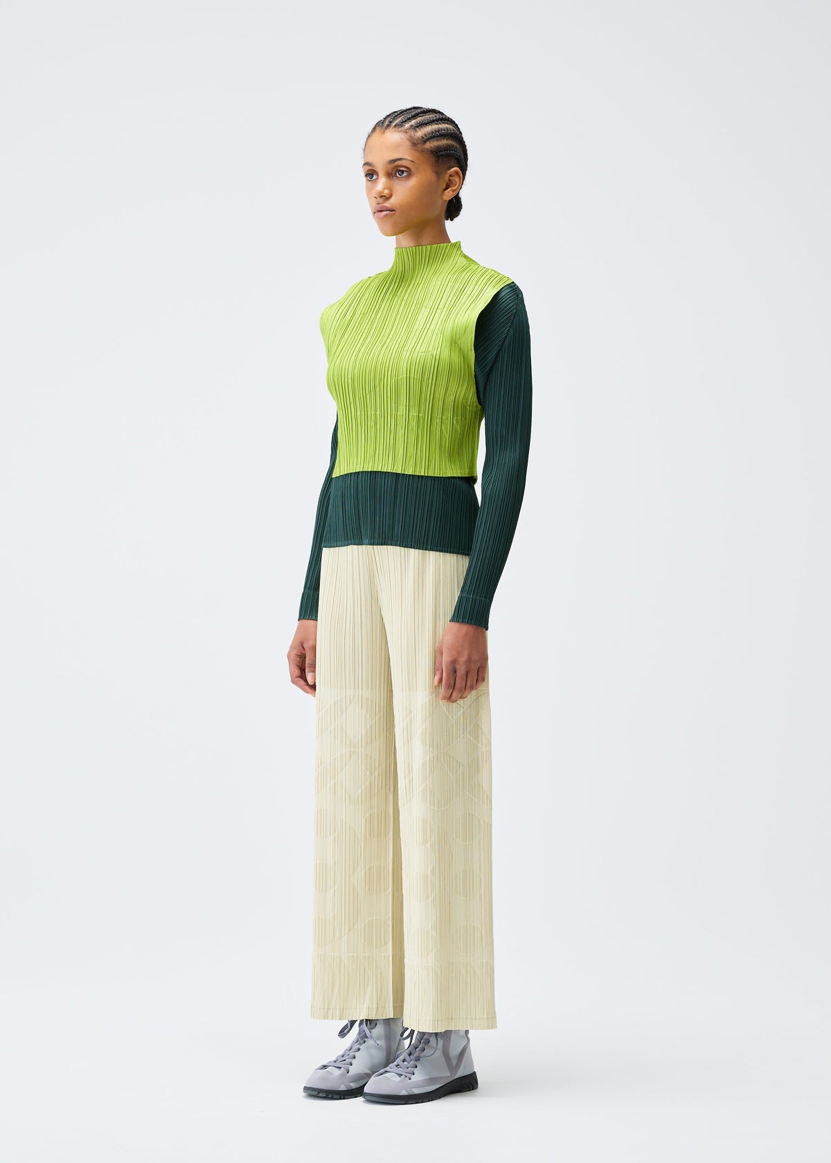 A person stands against a plain white background wearing the PLEATS PLEASE ISSEY MIYAKE TUNE: a green high-neck sleeveless top over a dark green long-sleeve shirt, wide-leg cream embossed pants, and gray lace-up shoes. view 6 of 8