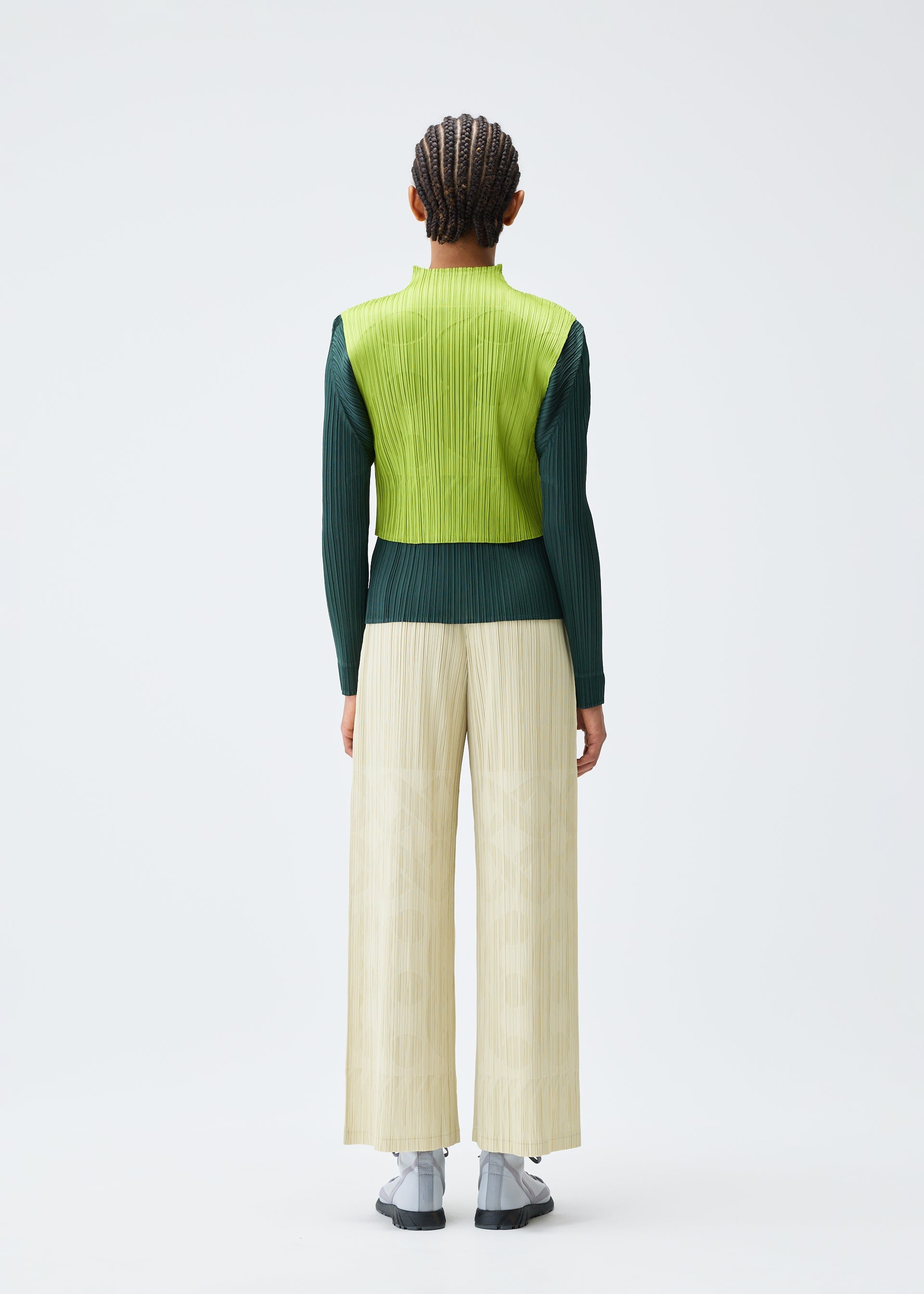 Zoomed view A person faces away, wearing the PLEATS PLEASE ISSEY MIYAKE TUNE lime green high-neck sleeveless vest over a dark green long-sleeve top, cream wide-leg pants, and white shoes, with tightly braided hair against a plain white background.