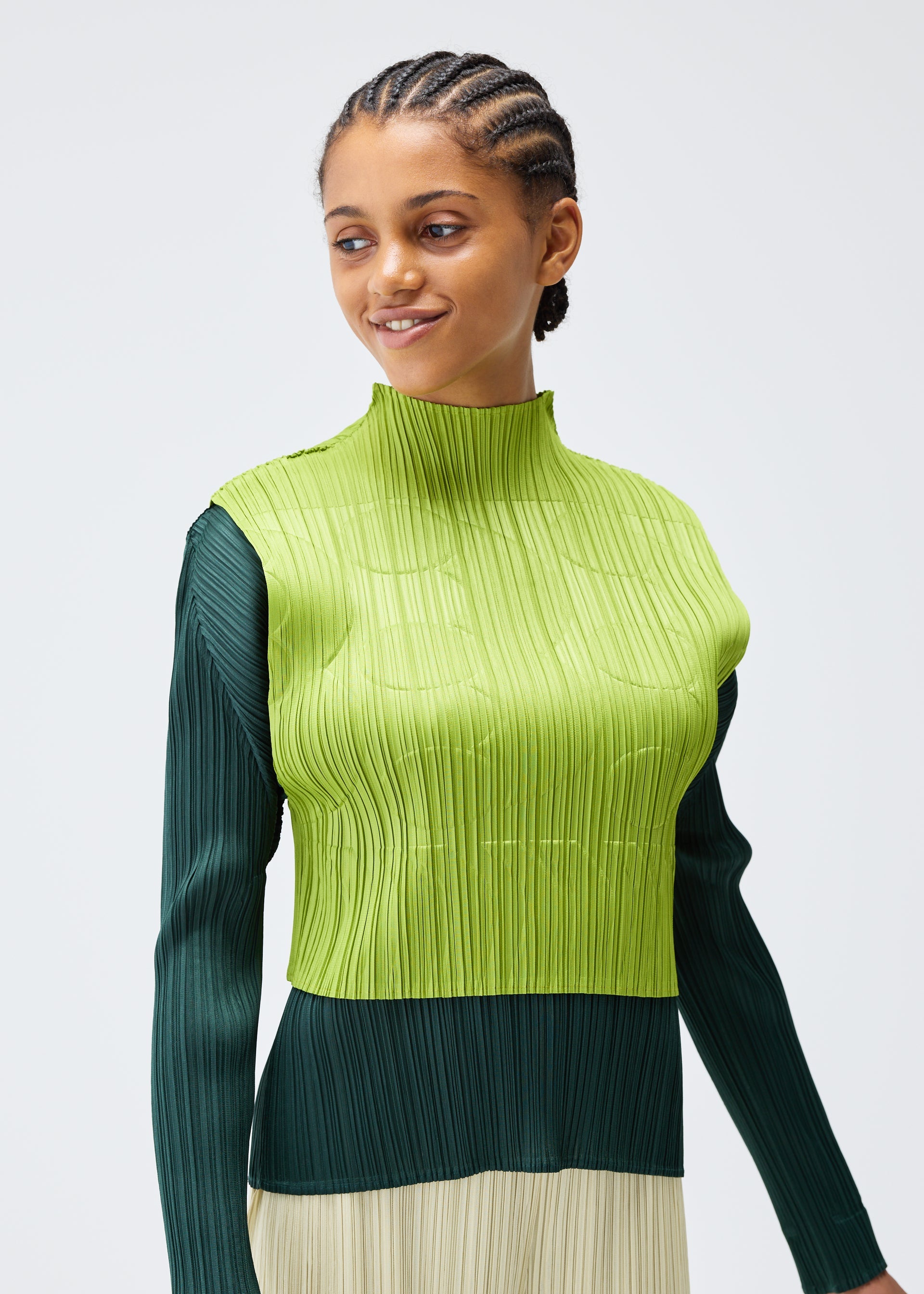 Zoomed view A woman with braided hair smiles softly, wearing the TUNE high-neck sleeveless top by PLEATS PLEASE ISSEY MIYAKE in lime green over a dark green long-sleeve shirt, creating a stylish layered look against a plain light background.