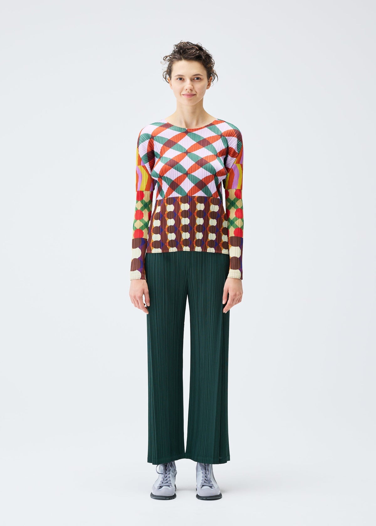 A person with short curly hair and a neutral expression stands facing forward against a white background, wearing the HARMONY top by PLEATS PLEASE ISSEY MIYAKE, dark green wide-leg pants, and light gray shoes. view 4 of 6