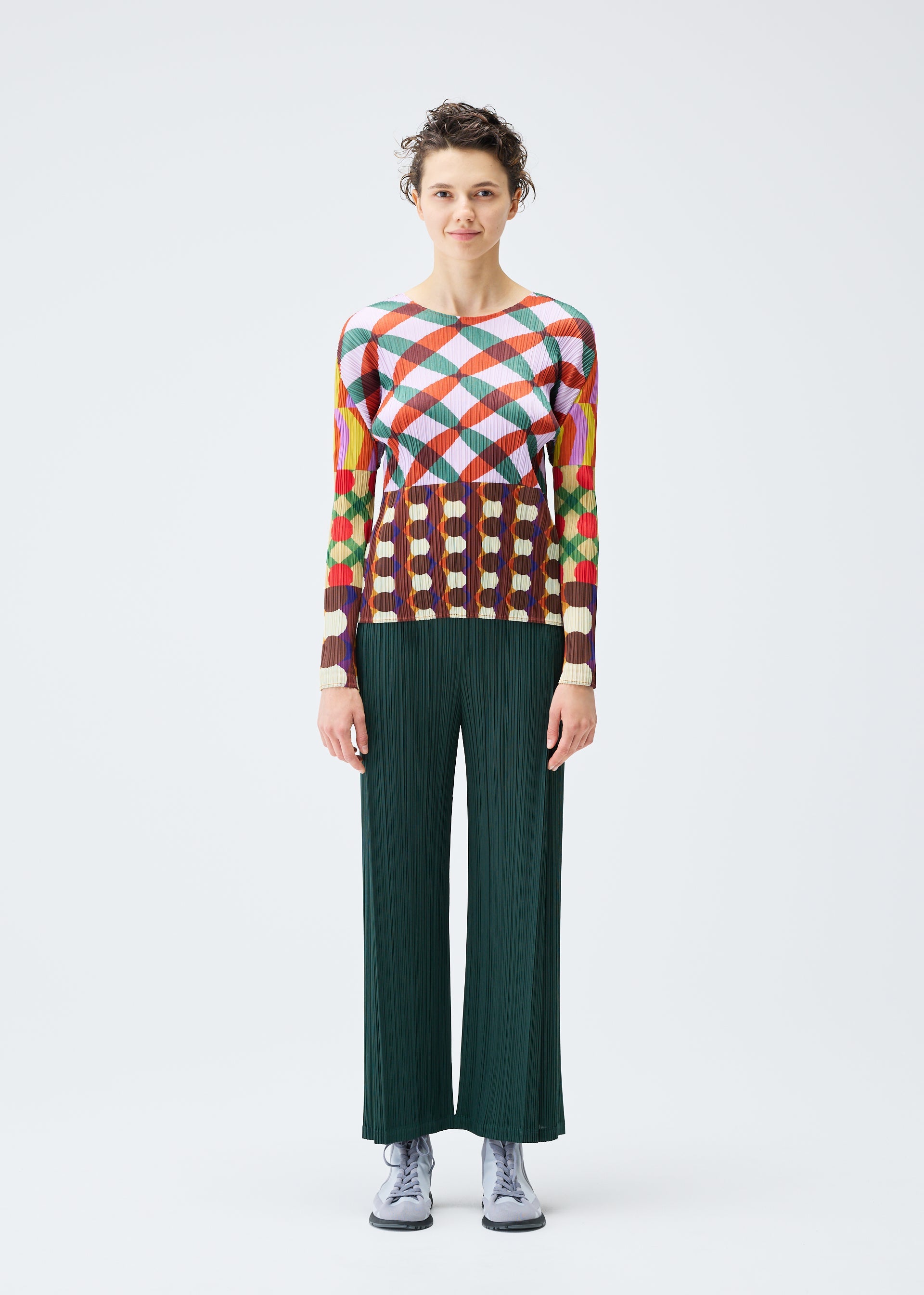 Zoomed view A person with short curly hair and a neutral expression stands facing forward against a white background, wearing the HARMONY top by PLEATS PLEASE ISSEY MIYAKE, dark green wide-leg pants, and light gray shoes.