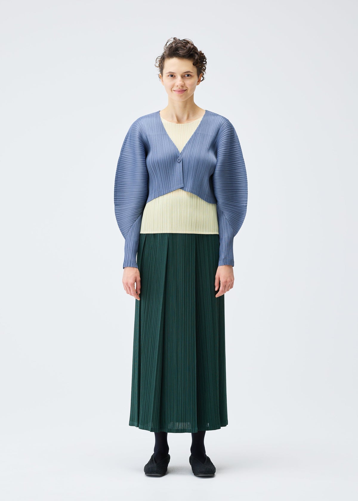 A person stands against a plain white background, wearing the PLEATS PLEASE ISSEY MIYAKE MONTHLY COLORS : DECEMBER textured light blue short cardigan over a pale yellow top and long dark green pleated skirt. They have short curly hair. view 7 of 9