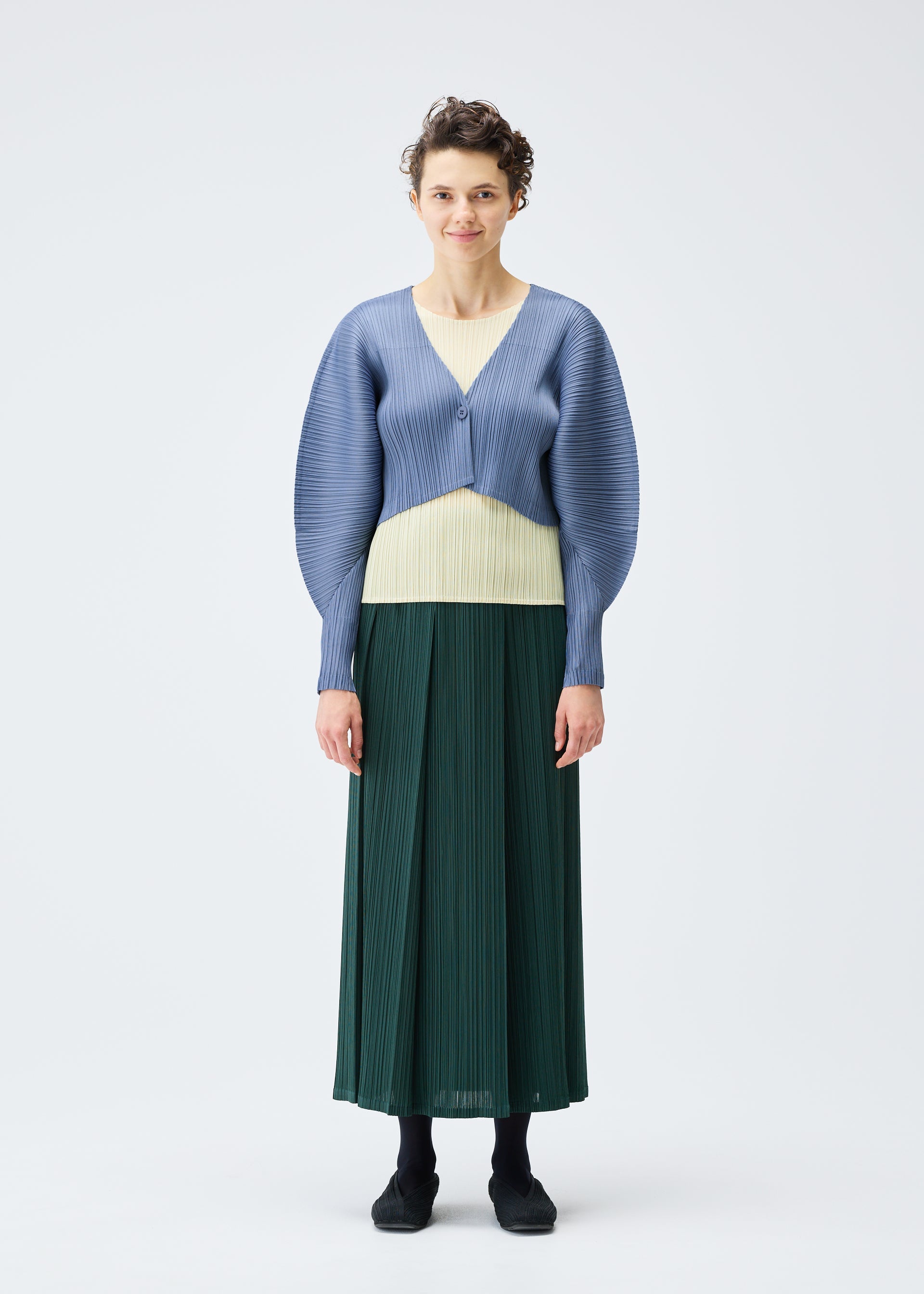 Zoomed view A person stands against a plain white background, wearing the PLEATS PLEASE ISSEY MIYAKE MONTHLY COLORS : DECEMBER textured light blue short cardigan over a pale yellow top and long dark green pleated skirt. They have short curly hair.