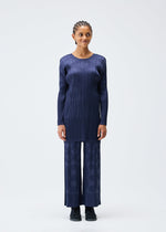 A woman with braided hair faces forward, wearing the TUNE matching set from PLEATS PLEASE ISSEY MIYAKE: a long-sleeved, round-neck tunic with vertical stripes and wide-leg pants with a subtle geometric pattern, paired with black shoes.