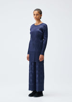 A person faces forward in the TUNE set by PLEATS PLEASE ISSEY MIYAKE—navy blue, long-sleeved pleated tunic with a subtle circular pattern and matching wide-leg pants. Braided hair and black shoes complete the look against a plain light background.