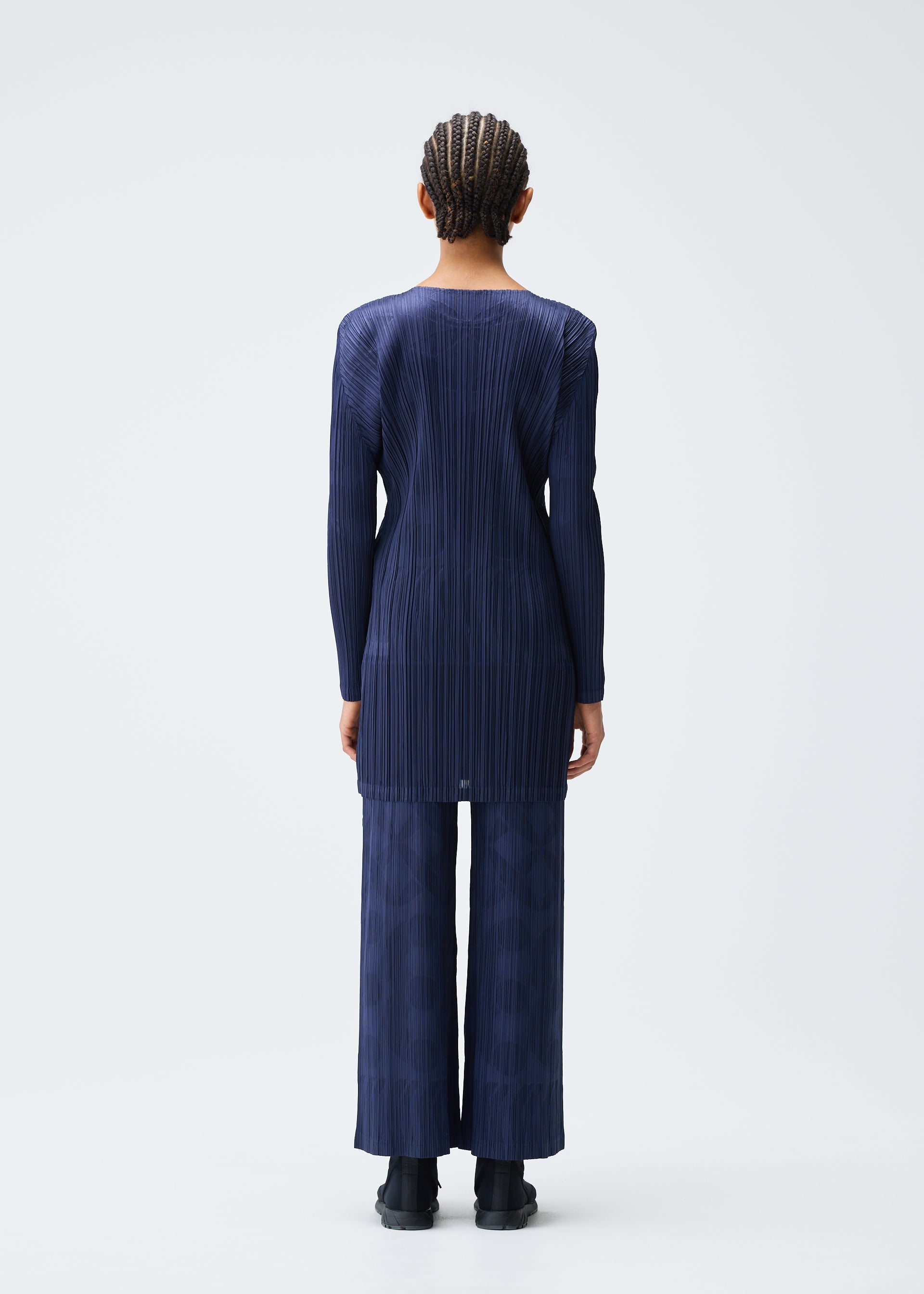 Zoomed view A person with braided hair faces away, wearing the TUNE long-sleeved navy tunic and matching wide-leg pants from PLEATS PLEASE ISSEY MIYAKE, paired with black shoes against a plain white background.