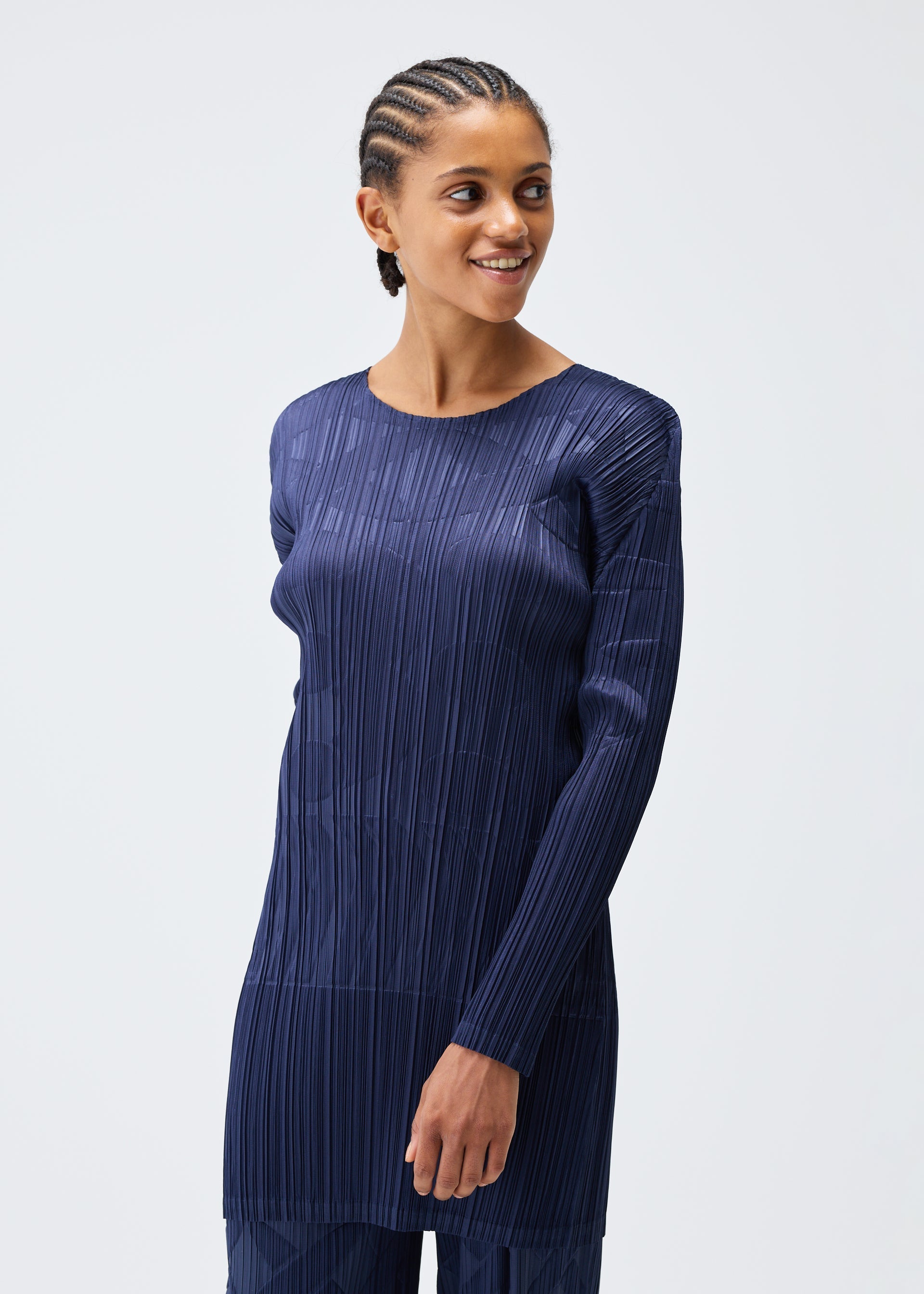 Zoomed view A person with cornrow braids smiles and looks to the side, wearing the dark blue TUNE round-neck tunic by PLEATS PLEASE ISSEY MIYAKE, featuring vertical pleats, against a plain light background.