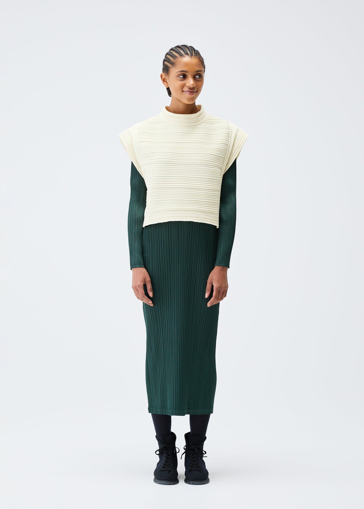 A woman with braided hair wears the PLEATS PLEASE ISSEY MIYAKE CREPE KNIT dress in dark green, layered with a cream textured vest, paired with black tights and sneakers, against a plain white background. view 4 of 6