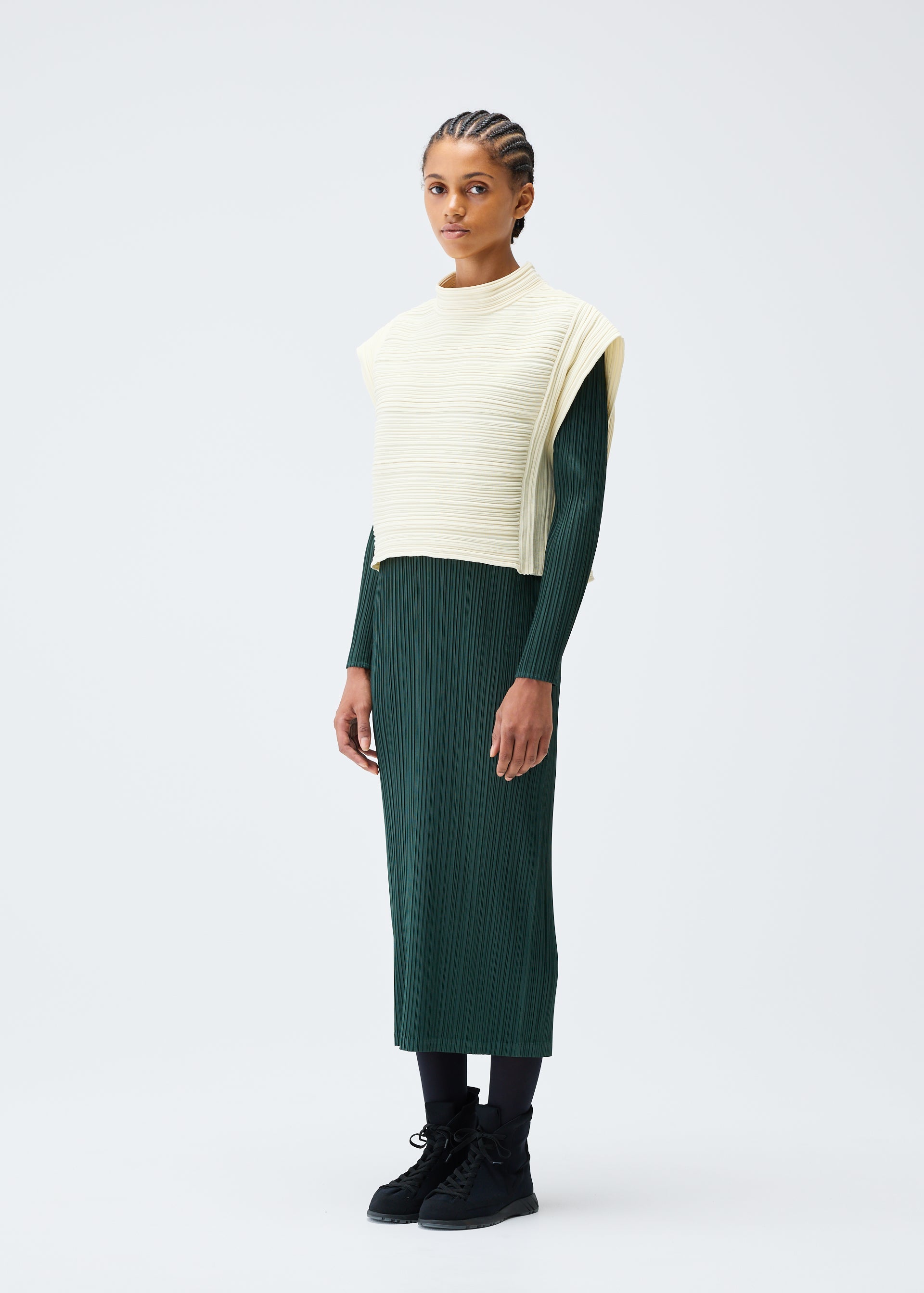 Zoomed view A person with braided hair stands against a plain background, wearing the PLEATS PLEASE ISSEY MIYAKE CREPE KNIT long dark green dress, black leggings and shoes, and a cream-colored lightweight vest.